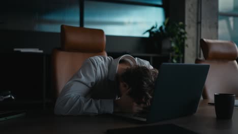 Sad-and-tired-office-worker-guy-with-curly-hair-puts-his-head-on-the-laptop-keyboard-and-shakes-it-to-the-side-during-a-difficult-task-in-the-office