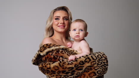 smiling blonde long-haired woman in fur coat holding a baby. mother kisses her beloved child on the head. grey backdrop.