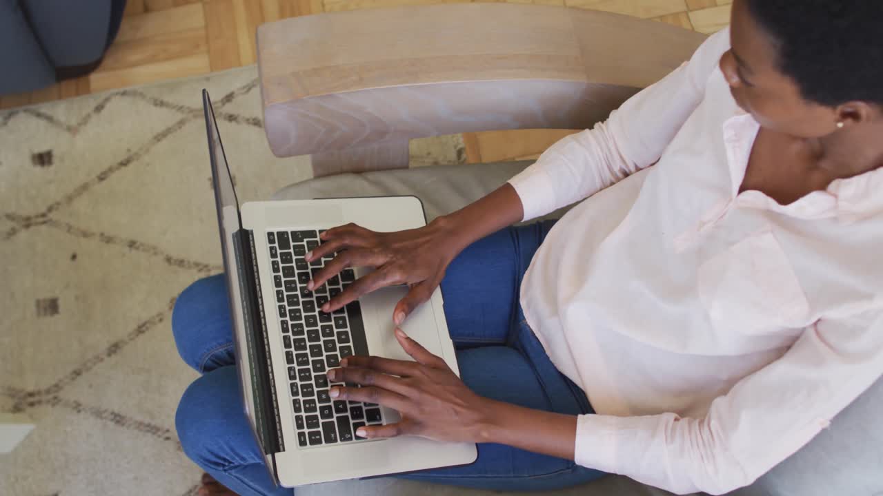 Happy african american woman sitting on armchair in living room, using laptop