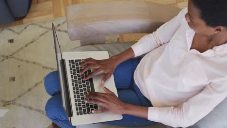 feliz mujer afroamericana sentada en un sillón en la sala de estar, usando una computadora portátil
