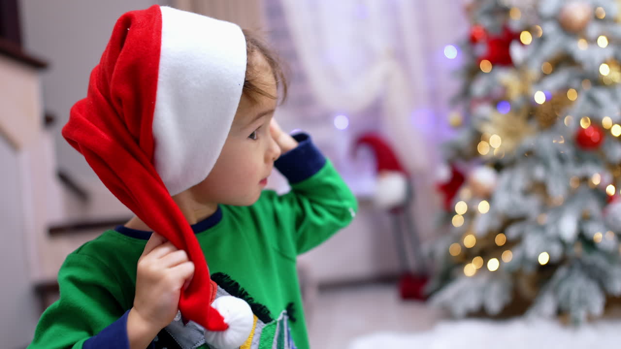 Beautiful toddler boy putting on red Santa hat on. Adorable baby celebrating Christmas. Close up.