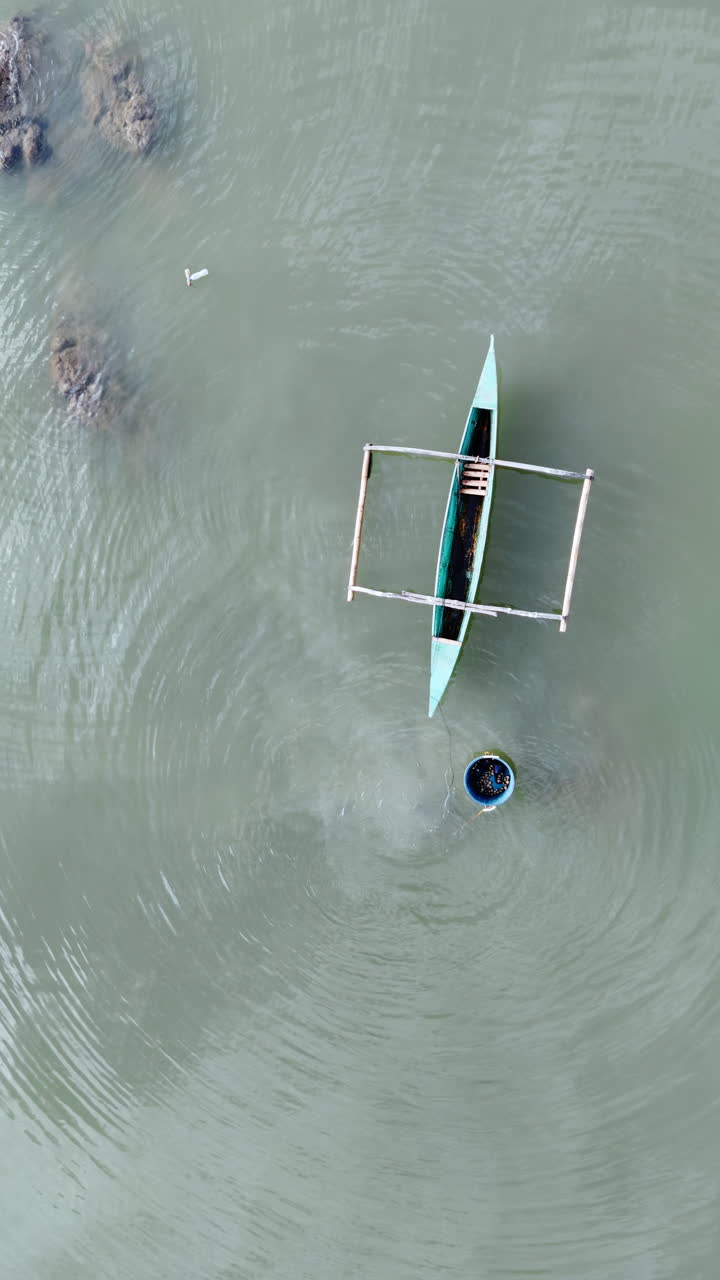 Aerial drone view of a man swimming near a traditional boat with a floating basket in shallow water in the Philippines. Vertical