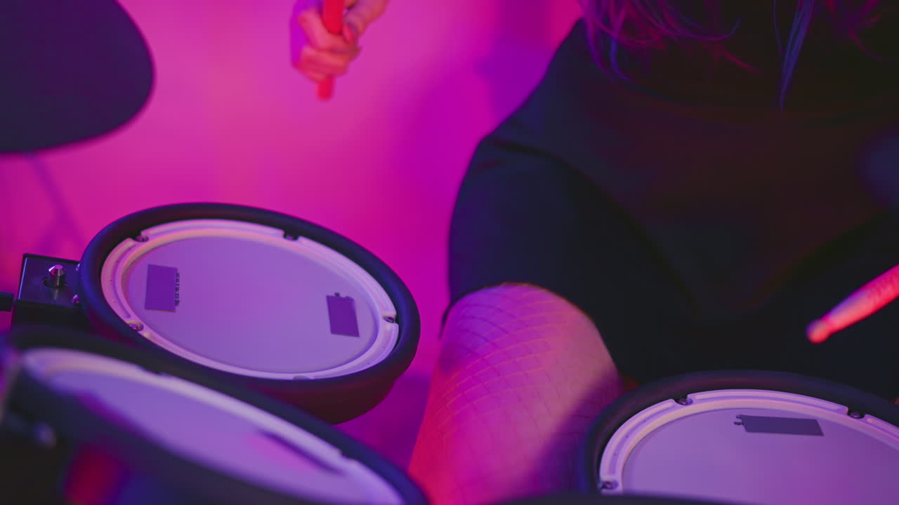 Partial close up of drummer legs in fishnet pantyhose seated at electronic drum kit, hands moving sticks to strike pads under pink and purple studio lighting, cables and module