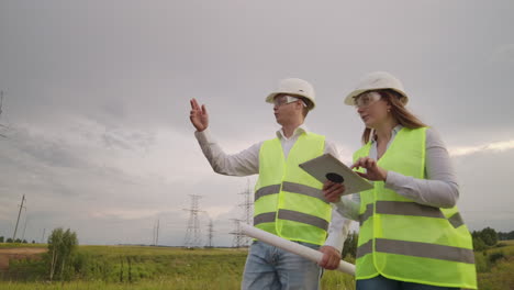 un electricista masculino y femenino en los campos cerca de la línea de transmisión de energía. es un electricista que gestiona el proceso de erección de líneas eléctricas. el mecánico con un casco y el gerente con una tableta