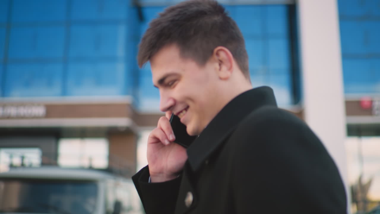 현대적인 유리 건물이 있는 활기찬 도시 지역에서 손으로 몸짓을 하면서 흥분하여 전화로 행복하게 이야기하는 검은 코트를 입은 은행가의 측면 시각