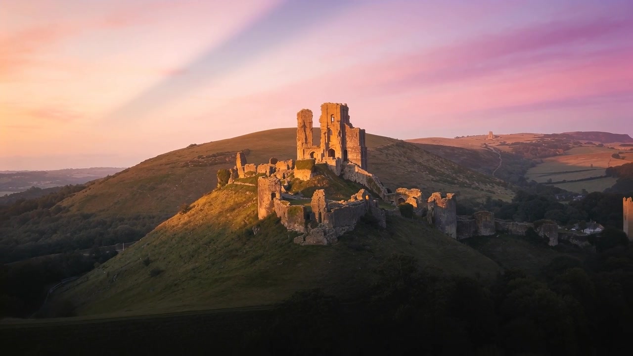 Drone in fase di lancio si avvicina a un castello di pietra su una collina, abbassandosi per rivelare dettagli e campi coltivati.