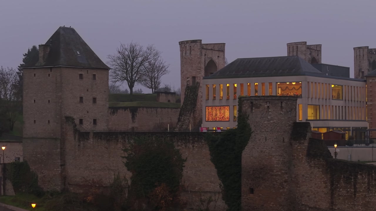 Lateral night drone shot revealing Tour Jacob in Luxembourg, surrounded by illuminated buildings and historic architecture, capturing the dramatic urban skyline and evening atmosphere.