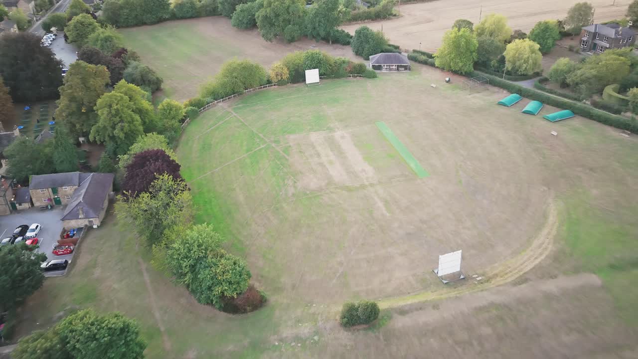 Descending drone motion, Wentworth’s cricket ground unfolds with its central pitch, white sight screens, green covers, and surrounding rooftops framed by trees and open countryside