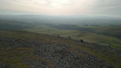 Hiker-on-rocky-hillside-with-long-orbit-reveal-of-patchwork-fields-and-mountain-Ingleborough-above-Ingleton-Yorkshire-UK