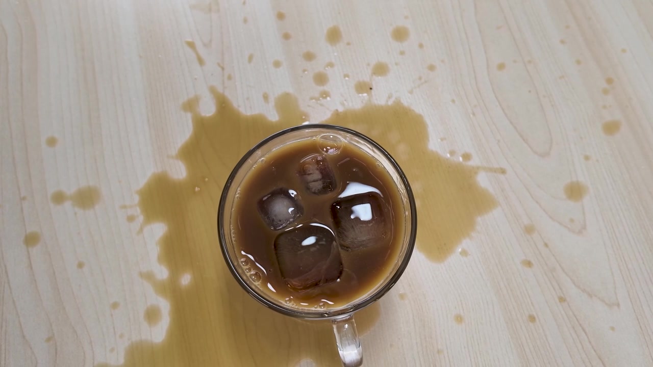 Overhead shot of spilled iced coffee surrounding a glass mug on a light wooden surface.