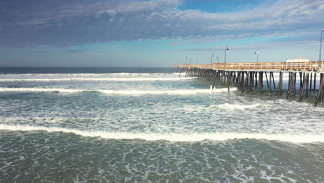 waves rolling into the beach alongside the landmark pier at pismo beach - aerial pull back