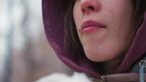 primer plano de una joven con mancha en la cara con capucha comiendo algo al aire libre, centrado en comer, comida casual, vibraciones de invierno, capucha, fondo borroso con árboles y atmósfera de invierno