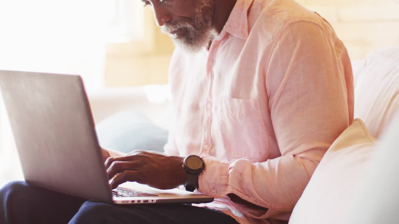 Happy senior african american man in log cabin, sitting on sofa and using laptop, slow motion