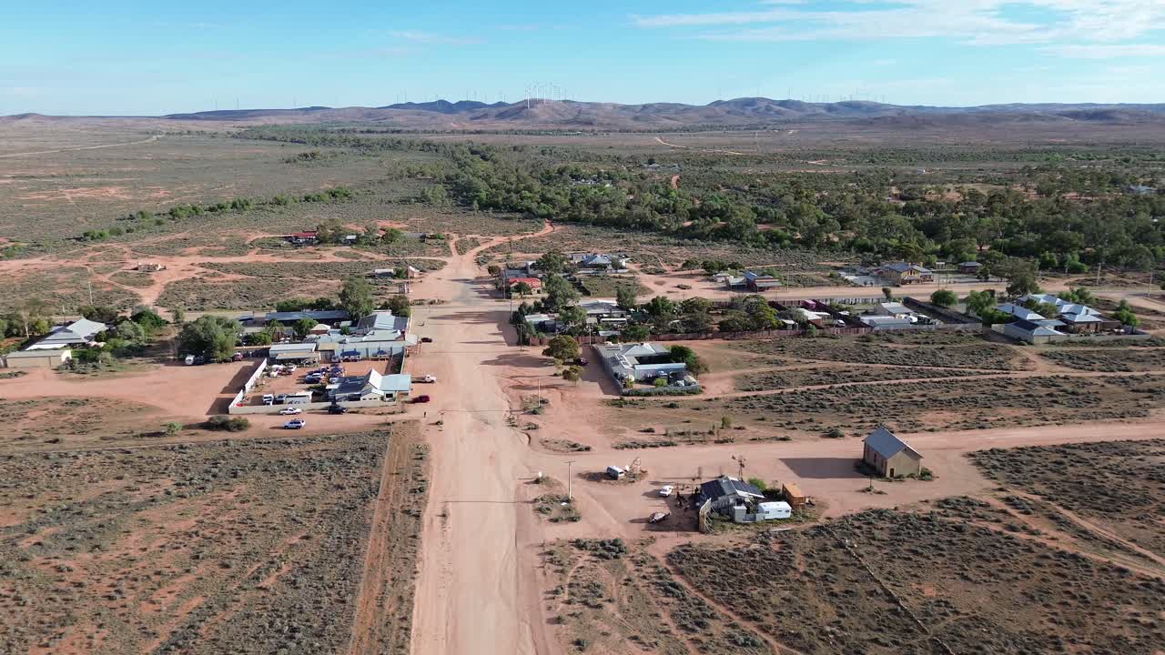las imágenes de drones se elevan sobre silverton en el interior de australia, mostrando un accidentado camino de tierra, vastos paisajes de tierra roja y turbinas eólicas girando contra el infinito horizonte.
