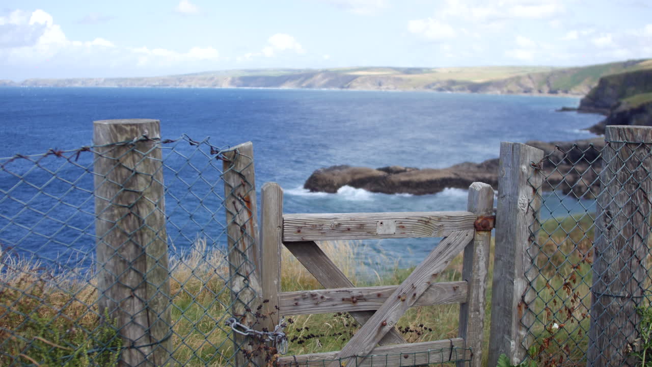A fence with a wooden gate is standing at the edge of a coastal path above the blue waters near Port Isaac and Portwenn, capturing Cornwall's wild English seascape and rocky shorelines