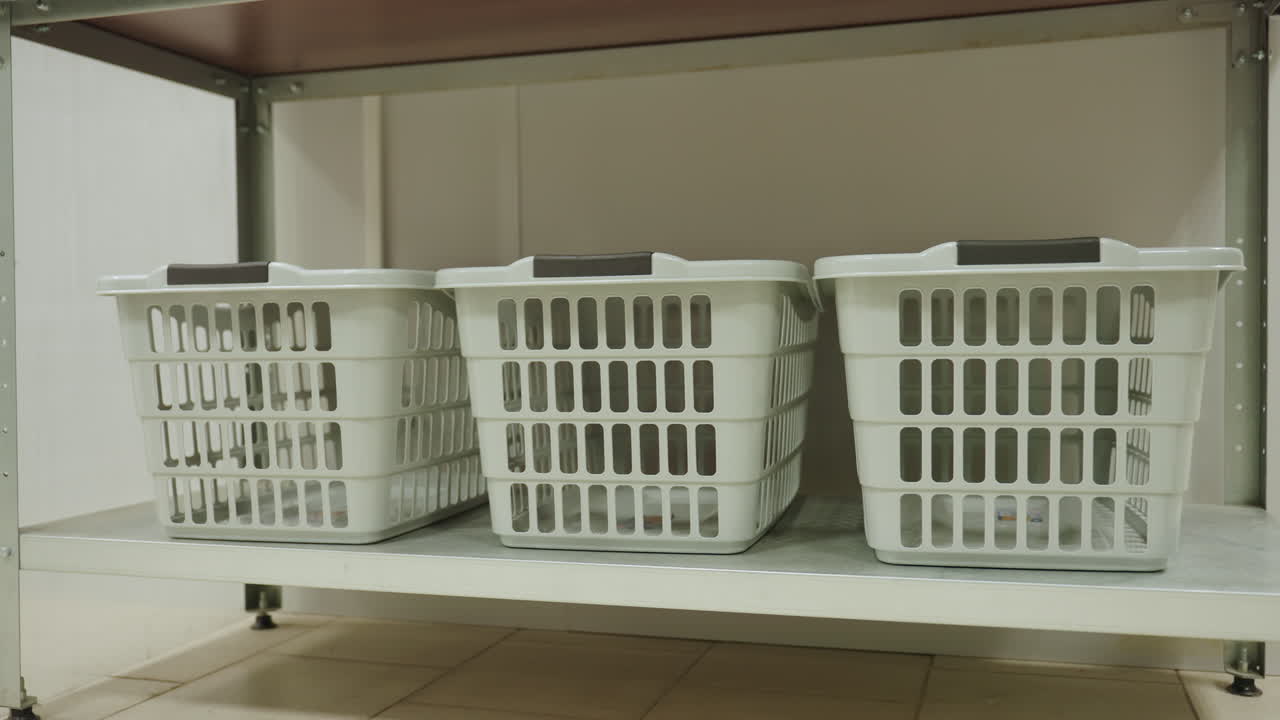 Three clothes hampers on metal shelf in tidy laundry room, empty plastic bins organized for service, clean utility storage scene with soft light, neutral tones, modern facility vibe