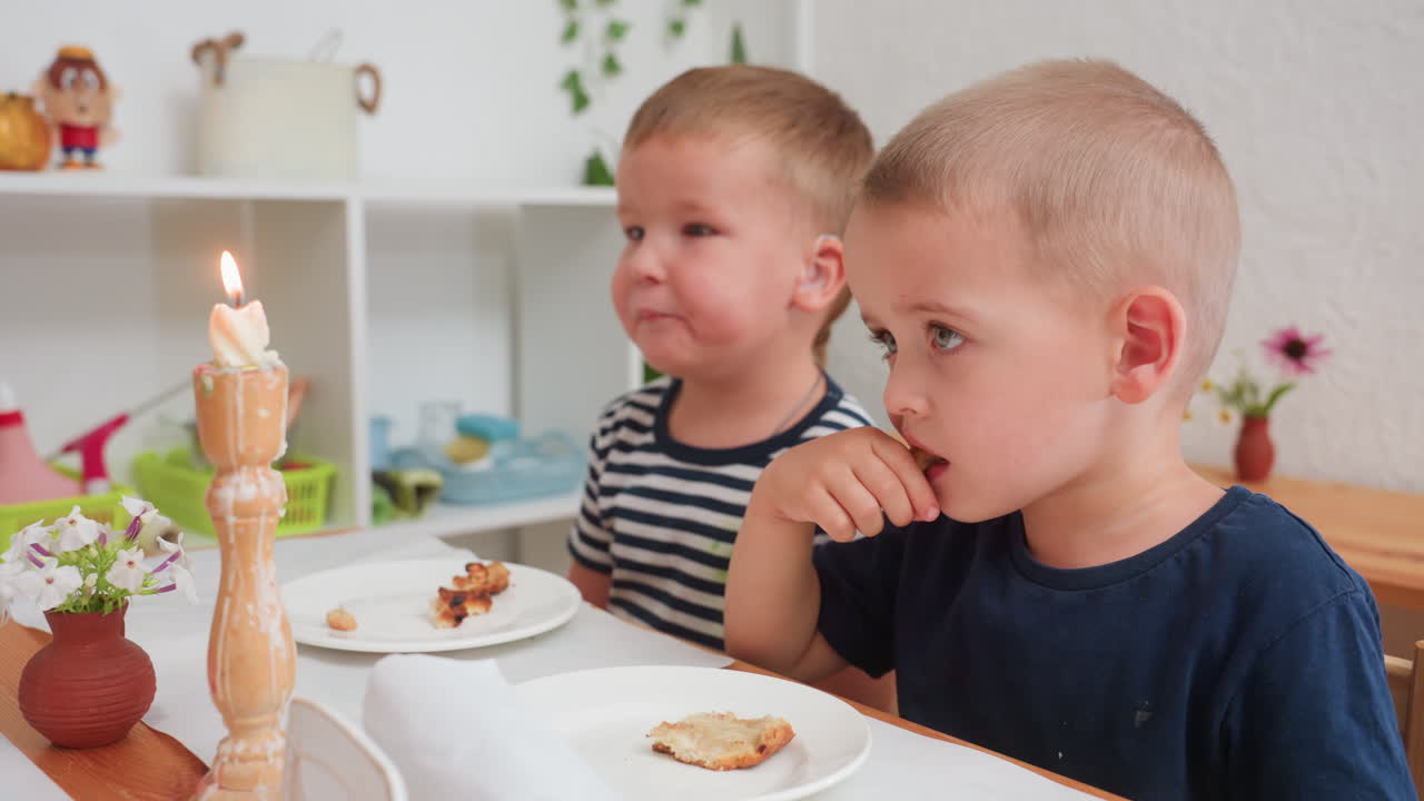 사시가 있는 남자아이가 촛불을 불어 끄려고 하지만 불꽃은 꺼지지 않고, 호기심 가득한 표정으로 남자아이들이 유치원 테이블에서 점심을 먹고 있어요. 접시에는 빵 부스러기가 있고, 따뜻한 교실 분위기예요.