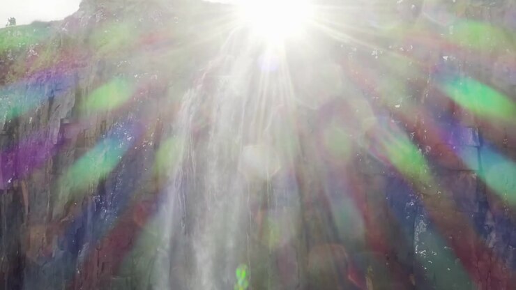 Aerial shot of a waterfall falling from a green rocky mountain and the sun casting light directly in the camera of the drone.