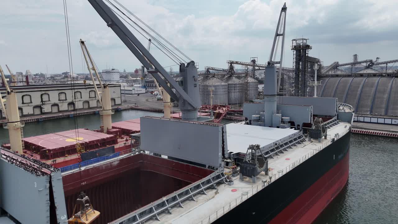A bulk carrier is loaded with grain at a port terminal. Cranes and conveyor systems handle the cargo efficiently, with silos and storage facilities visible in the background.