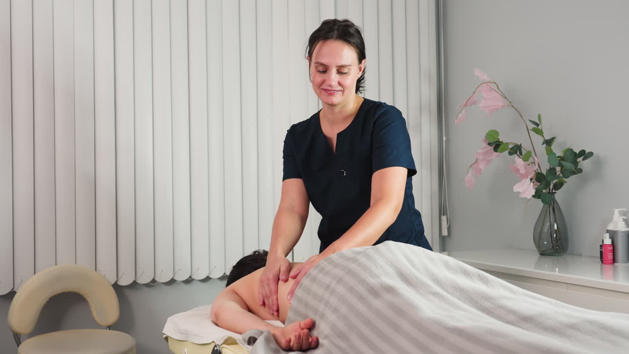 Cheerful professional smiling while rubbing back of female client on massage bed in spa room with flower vase and cream on shelf under soft lighting creating relaxing wellness moment