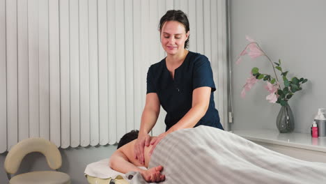 cheerful professional smiling while rubbing back of female client on massage bed in spa room with flower vase and cream on shelf under soft lighting creating relaxing wellness moment