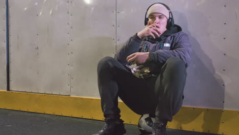 young man wearing headphones seated on ball resting against metallic wall with yellow paint, lighting cigarette and exhaling smoke in relaxed posture