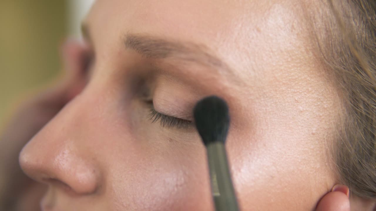 Extremely close of a young woman's face. Make up artist putting on nude eyeshadows using two brushes at the same time