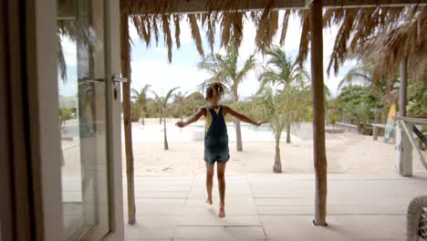 Caucasian-girl-running-and-raising-hands-at-swimming-pool-at-beach-house-with-palm-trees