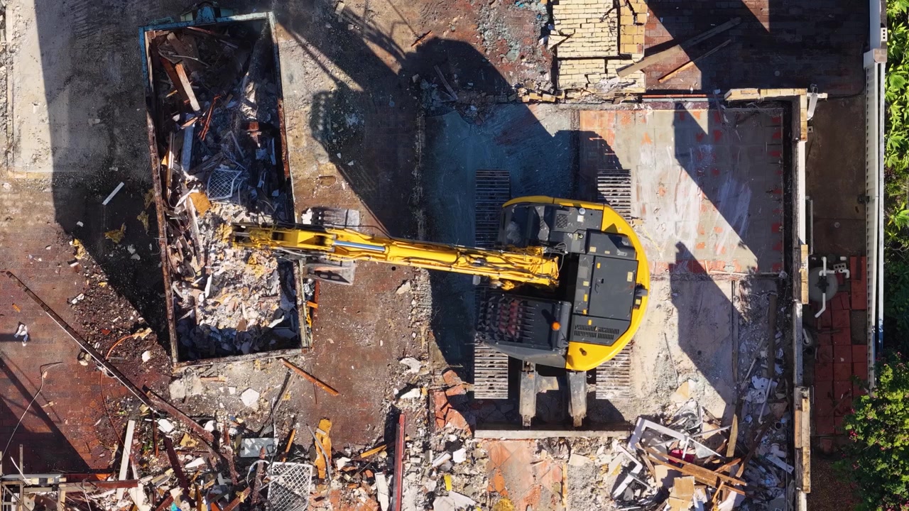 Droneopptak ovenfra viser en gul gravemaskin som laster bygningsavfall i en avfallscontainer i sollys.