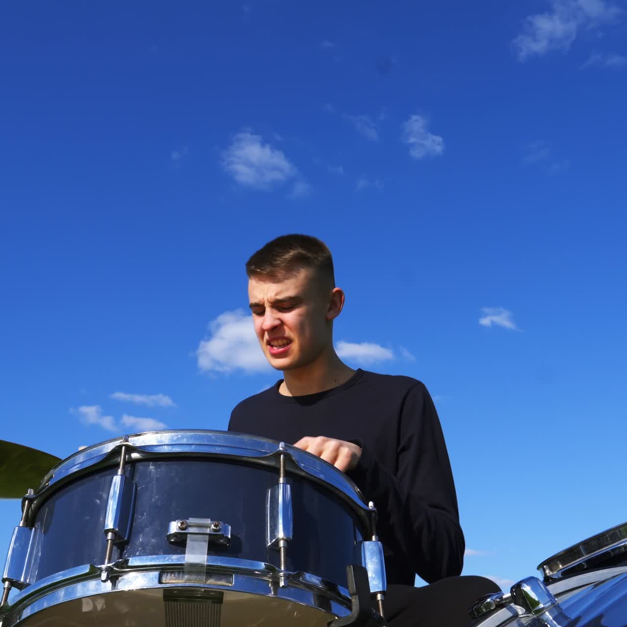 Young Caucasian man playing drums savagely. Emotional male musician hitting drums and cymbals with wooden sticks at blue sky backdrop. Low angle view