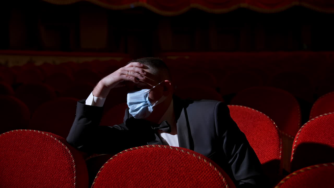 Theater hall after pandemic. Male in medical mask sits alone in empty hall with red seats. Covid-19 concept.