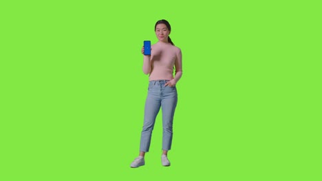 초록색 스크린을 맞은 카메라를 향해 파란색 화면 휴대전화를 들고 있는 웃는 여성의 전체 길이의 스튜디오 초상화 1
