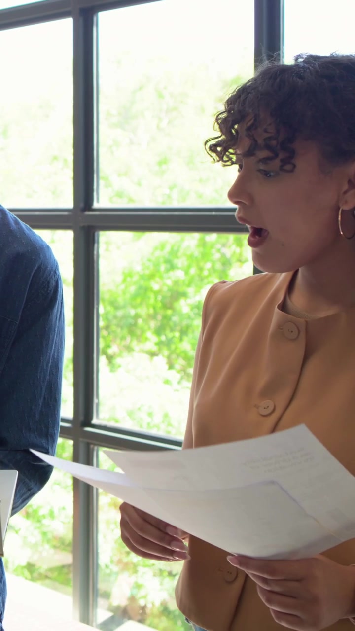Video verticale: Donna che legge con un gilet beige, tiene due fogli vicino a una finestra e guarda il laptop.