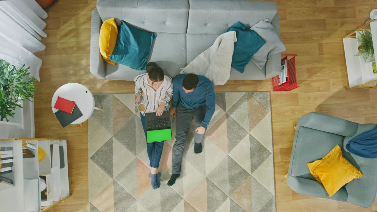 Young Couple is Sitting on a Floor and Using a Laptop with Green Screen. Girl Drinks Coffee. Cozy Living Room with Modern Interior with Carpet, Sofa, Chair, Table, Shelf, Plant and Wooden Floor. Top Down.