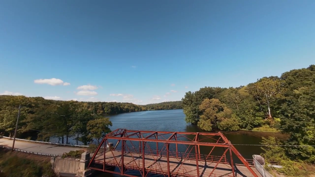 Vista aérea FPV de uma ponte de aço abandonada sobre o reservatório Muscoot em Westchester, NY. Estrutura histórica sobre a água com árvores ao redor em um dia ensolarado. A câmera faz um movimento de dolly in com movimentos dinâmicos