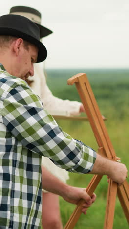 a woman in a white dress, holding a painting brush, watches her husband who is wearing a black hat and plaid shirt as he focuses on setting up a wooden tripod in a scenic grassy field