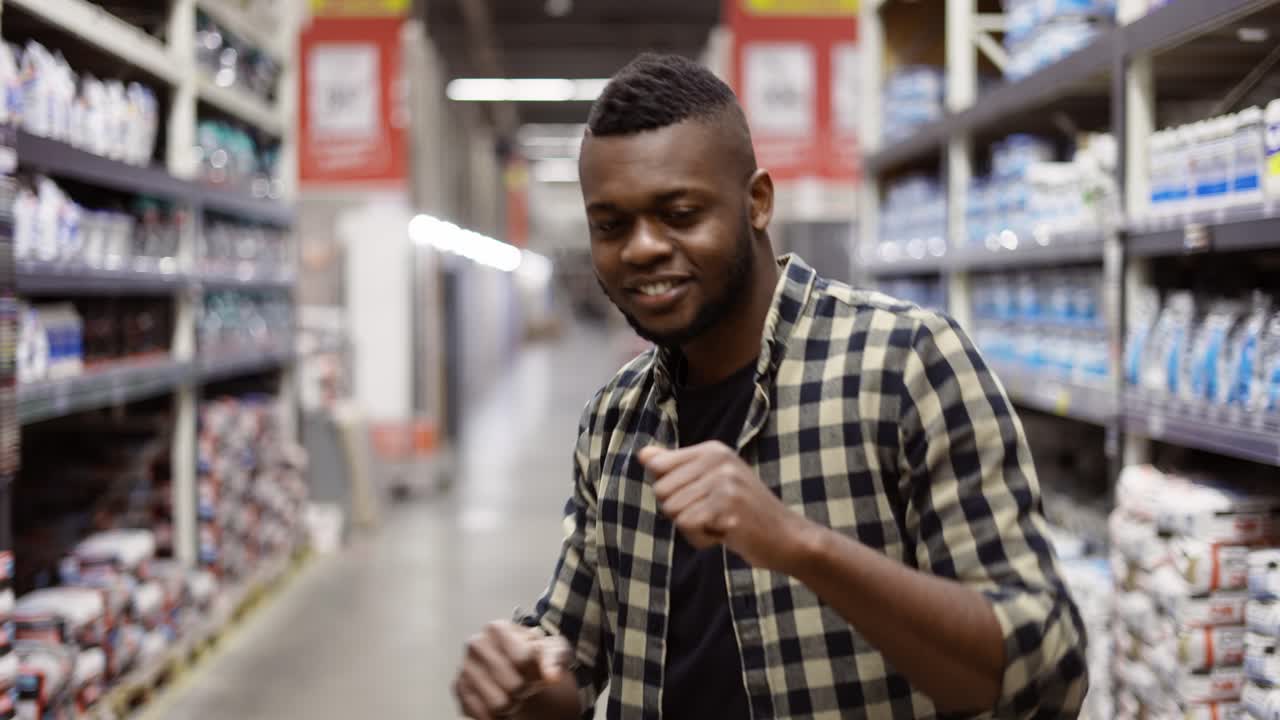 Smiling guy dances gesturing in hardware goods store between stacks slow motion