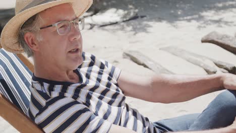 happy senior caucasian man sitting in deckchair talking on sunny beach, in slow motion