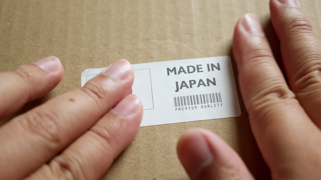 Hands applying MADE IN JAPAN flag label on a shipping box with product premium quality barcode