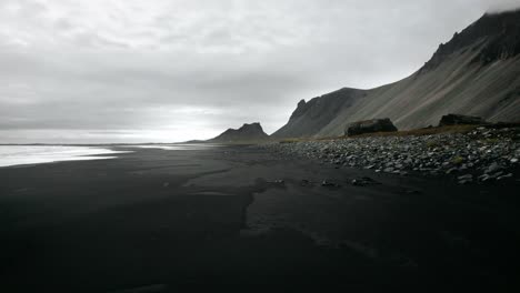 Flying-through-rock-formation-on-black-sand-beach-stokksnes,-volcanic-mountain-landscape