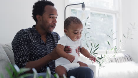 Father-Sitting-On-Sofa-At-Home-Playing-Game-Bouncing-Baby-Son-On-Knee