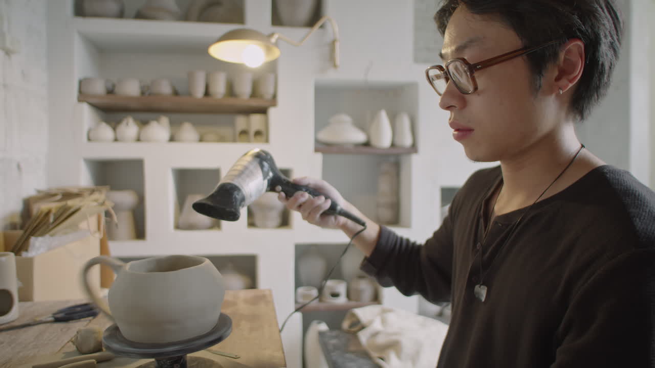 Young Ceramist Drying Clay Mug on Pottery Wheel with Hairdryer