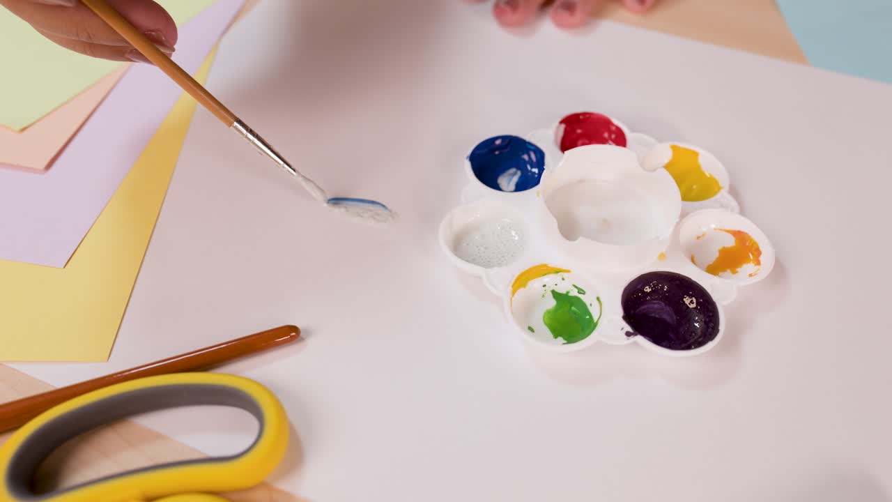 Person mixes watercolor paints with brush on palette, bright workspace, overhead view, soft lighting