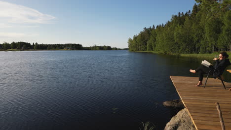 A-woman-sitting-on-deck-on-the-coast-of-Finland,-drinking-wine-and-reading-a-book