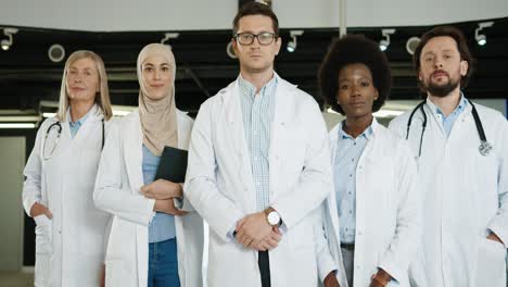 Multiethnic-doctors-team-in-medical-coats-standing-in-hospital-lab-and-looking-at-camera