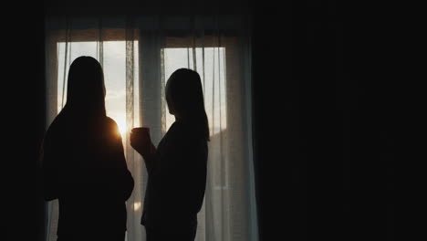 A-woman-with-a-teenage-daughter-stand-at-the-window-at-sunset.-Holding-cups-of-tea.-Rear-view