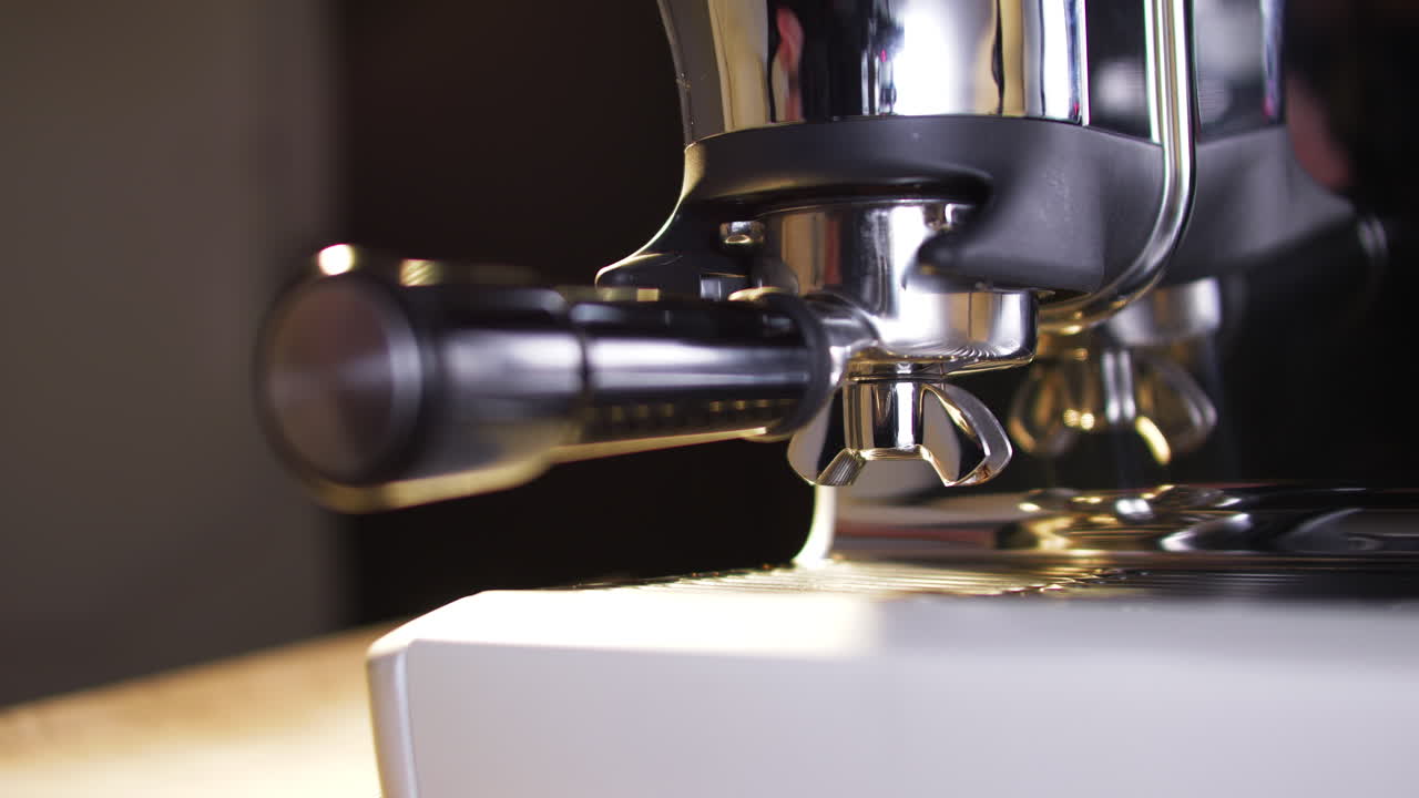 Um close-up mostra uma mão soltando e removendo um portafiltro de um moedor de café expresso cromado. O metal polido e a iluminação suave destacam o fluxo de trabalho diário do barista e a preparação do café.