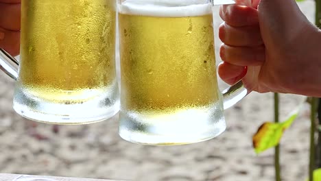 close-up of two hands clinking frosty beer mugs with a sandy beach in the background.
