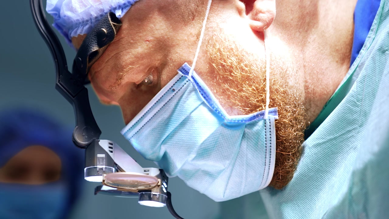 Volto di un medico caucasico adulto barbuto con mascherina, cuffia e loupes chirurgiche. Ritratto ravvicinato di un chirurgo concentrato sull'operazione