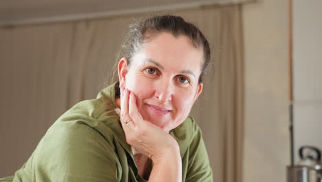 portrait view of white woman in green dress and white apron gently resting head on hand with soft smile while glancing at camera in peaceful moment, radiating calm and warmth